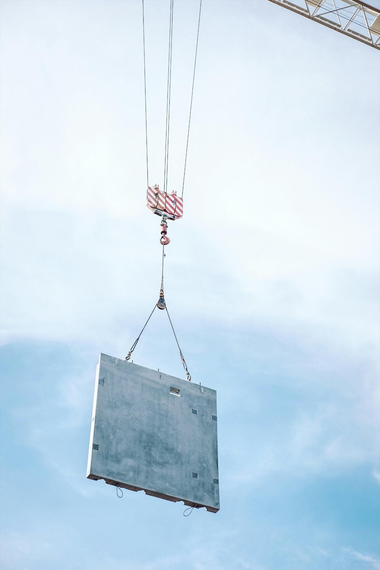 A concrete panel suspended from a crane against a clear sky.